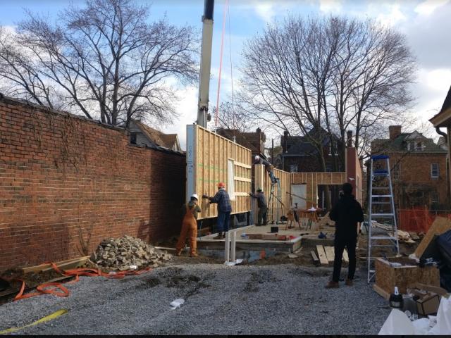 The walls are built in a factory under controlled conditions. Continuous laminated veneer lumber at roof framing acts as header. Furring strips form an interior wiring chase behind the drywall that minimizes penetrations in the thermal envelope.